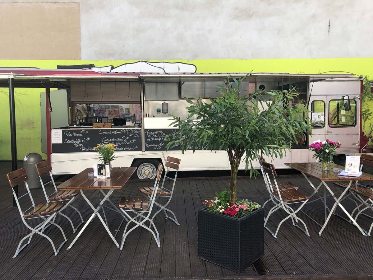 Weiße Bistrotische und Klappstühle vor einem Foodtruck mit Tafelmenü im Biergarten des Ballhaus Berlin