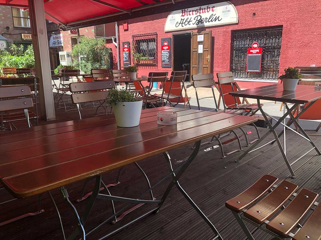 Holztische und Klappstühle auf der überdachten Terrasse vor der roten Fassade der Bierstube Alt Berlin